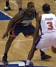 A black person wearing a dark-color jersey defends against another person in white jersey who dribbles in front of him