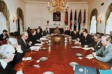 Thatcher is the only woman in a room, where a dozen men in suits sit around an oval table. Reagan and Thatcher sit opposite each other in the middle of the long axis of the table. The room is decorated in white, with drapes, a gold chandelier and a portrait of Lincoln.