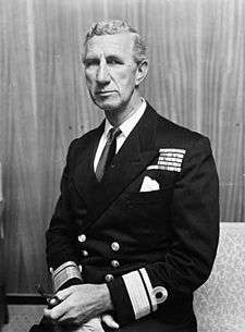 Portrait of a man in naval uniform. Visible from the waist up, he appears to be sitting and wears a dark, formal naval jacket that sports the rank of rear admiral. There are medal ribbons on his left breast, and he is holding a smoking pipe in his left hand.