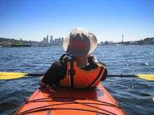 Photo of rear of person wearing orange life preserver sitting in kayak with buildings in far background