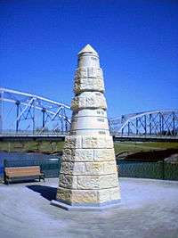 A stone obelisk indicating the level of past floods at different heights. In the background a bridge and a river can be seen.