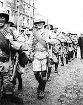 Soldiers in colonial-style military uniforms march past the camera, smiling