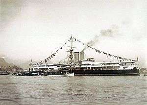 A photograph showing a steamship in port, fully dressed with flags and a single visible gun turret towards the bow