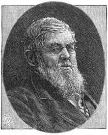 A bespectacled man with gray hair and a long beard wearing a black jacket and white shirt