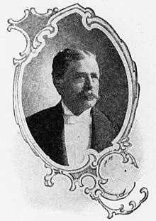 Head and shoulders of a white man with mustache wearing a tuxedo and white bow tie. The portrait is surrounded by a decorative oval-shaped frame.