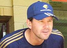 A white man with stubble, wearing a dark blue baseball cap with three white stripes on the peak and a yellow logo on the front.  He is wearing a dark blue top with three yellow stripes down each arm from the shoulder and is leaning forward in front of a doorway.