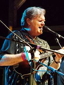 A man wearing a black shirt and playing a stringed instrument with his fingers. His eyes are closed, and he is standing behind microphone stands.