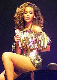 A young brunette woman sitting on a piano. She wears a golden dress and holds a microphone in her right hand
