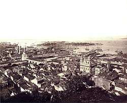 Photographic aerial view of the city from a hillside with ships at anchor in the harbor and island in the background