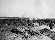 Soldiers march along a sunken dirt road in small groups. Either side of the road debris is strewn and the ground has been churned up from recent artillery attacks