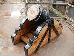 A squat black mortar, the end gapped with a wooden plug on which is carved "Roaring Meg"; the mortar has wooden supports with black metal brackets.