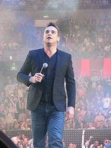 A middle-aged, brown-haired man wearing a jacket and jeans walks on what appears to be a stage, holding a microphone to his chest and looking upward. An audience is in the background.