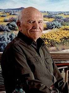 A man of around 90 sits in front of a landscape painting of desert wildflowers