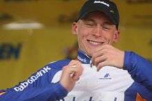 A smiling man in his mid twenties wearing a white and blue cycling jersey and a black baseball cap.