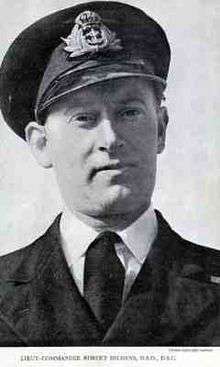 Head and shoulders picture of Robert Peverell Hichens in Royal Navy dress uniform of peaked hat, buttoned tunic, white shirt and black tie. Just visible of the right breast is the medal ribbon of the Distinguished Service Cross