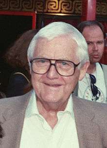 Photograph of a man's head. He is smiling slightly, has white hair, and is wearing glasses. He is wearing a sports jacket and a white shirt with an open collar.