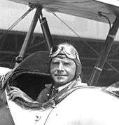 Photograph of Roderick Burnham seated in a biplane, looking backwards toward the camera, and wearing a period pilot's cap and goggles.