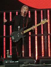 Roger Waters, dressed in black, playing a bass guitar and speaking into a microphone. Behind him are several red vertical video panels.