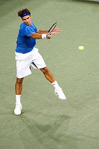 A brown-haired male tennis player with white shorts, a blue shirt and a blue headband swings a right-handed forehand on a hard court surface