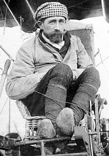 A black and white photograph of Roger Sommer, a man with a short beard, sitting in a Farman biplane seat surrounded by wires