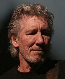 Roger Waters playing a bass guitar and singing into a microphone. He has grey hair and is unshaven.