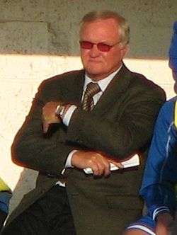 A man sitting in a dugout with his arms folded during a football match. He is wearing a pair of sunglasses as he is looking into the sun.