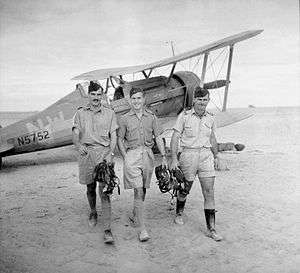 Three men in khaki shirts and shorts with forage caps, in front of a biplane