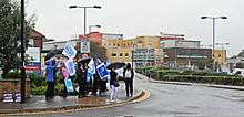A group with umbrellas and holding signs with slogans saying "RCN says..." stand on the corner of a road, in front of a hospital