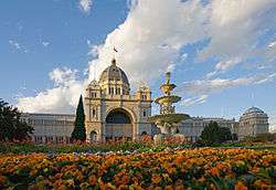 Large cream-coloured building with central dome and grand arched entrance, fronted by flowered gardens and a tiered fountain