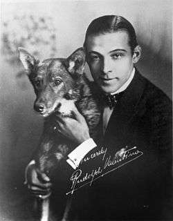 A black and white photograph of Rudolph Valentino with his dog; Valentino wears a dark suit and bow tie and is shown from the waist up, looking at the camera. The photograph has his autograph in white.