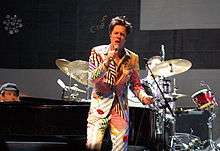 A man wearing a striped multi-colored suit with his eyes closed, singing into a microphone on a stage. Behind him is a man sitting behind a drum set and a man sitting at a piano.