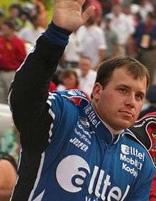 A man in his early thirties wearing a blue jacket with sponsors' logos. His right arm and hand are elevated in a waving gesture.