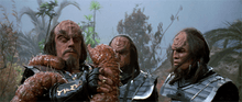 Two aliens watch their leader fight a slimy worm that has wrapped itself around his arm and neck. The aliens are wearing metallic armor with decorative script on their gauntlets; their hair is long and black, and their foreheads are bumpy and ridged.