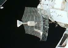 A rectangular dish shape of scaffolding covered in transparent sheeting, with a white insulation-covered radio receiver and support projecting from the centre. The blackness of space serves as the backdrop.
