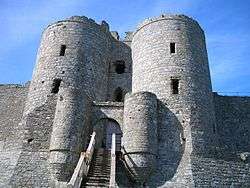 Two cylindrical stone towers flanking a gateway, and behind them two larger cylindrical towers. A path leads up to the gateway and curtain walls are attached to the towers.