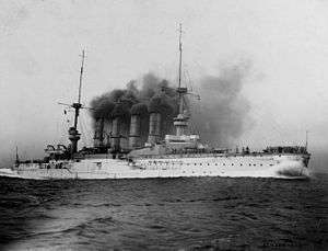 A large white warship belching thick black smoke plows through the water