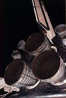 Three bell-shaped rocket engine nozzles projecting from the aft structure of a Space Shuttle orbiter. The cluster is arranged triangularly, with one engine at the top and two below. Two smaller nozzles are visible to the left and right of the top engine, and the orbiter's tail fin projects upwards toward the top of the image. In the background is the night sky and items of purging equipment.