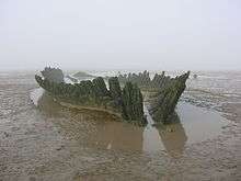Wooden hulk of ship, surrounded by wet sand.