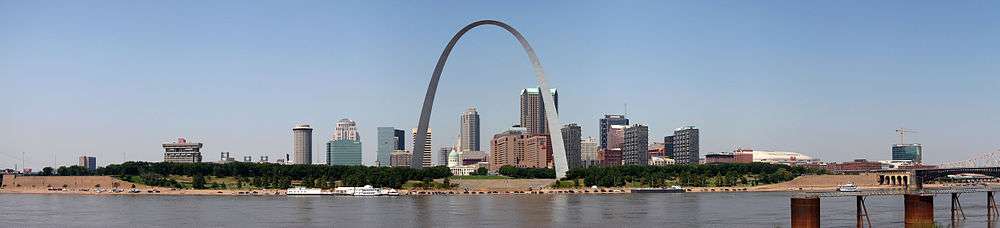 A large arch is in the center, across from a river. A clump of tall buildings is scattered behind it.
