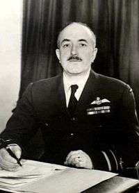 Half-length portrait of mustachioed man in military uniform with pilot's wings on left breast pocket, seated at desk with pen and papers