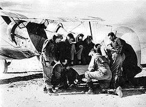 Half a dozen or so men looking at a map on the tail unit of a military biplane