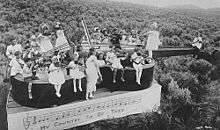 Black and white image of a group of children holding stringed instruments, sitting and standing in various position on a violin-shaped float in an open field.