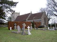 Gray stone building with red tiled roof, partially obscured by a hedge. A square tower is at the far end. The foreground includes several crosses and gravestones.