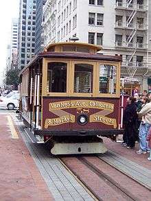 A cable car with the words "Van Ness Avenue, California, and Market Streets" on the front is stopped next to people who are about to get on.