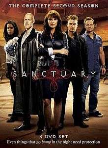 A DVD boxset cover consisting of five people standing behind a logo saying "Sanctuary". In the front is a middle-aged brunette woman wearing a black leather jacket and she is crossing her arms. To her right is a bald man with a long black leather overcoat, and a young woman of Indian descent. To the left are two men with short hear, one wearing a black jacket and jeans, the other with a blue shirt and jeans. The background indicates they are inside a large room.