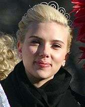 A close up of a smiling young woman, her blonde hair pulled back, wearing a silver tiara, silver earrings and a black cowl-necked coat. She is standing outside in bright sunlight.