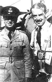 Half-portrait of two men, one wearing light-coloured uniform with dark peaked cap and pilot's wings on left-breast pocket, the other with moustache and wearing civilian clothes and parachute harness
