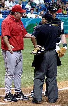 A man in a red warmup with gray pants stands arguing with a man in a black baseball jersey and dark gray pants.