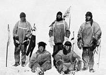 Five men(three standing, two sitting on the icy ground) in heavy polar clothing. All look unhappy. The standing men are carrying flagstaffs and a Union flag flies from a mast in the background. Scott's party at the South Pole.  Left to right: Oates; Bowers; Scott; Wilson; Evans