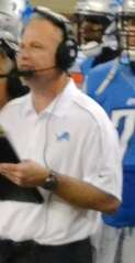 Linehan wearing a white Detroit Lions polo shirt and a headset and holding a clipboard on the Lions sideline